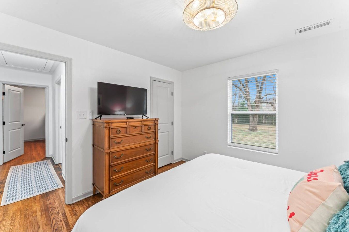 Un dormitorio con cama, cómoda, televisor encima de la cómoda, ventana con vista al exterior y puerta que da a otra habitación.