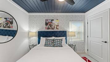 Une chambre à coucher avec une tête de lit capitonnée, un miroir rond, deux tables de chevet avec des lampes, un grand lit avec des coussins à motifs et une porte.