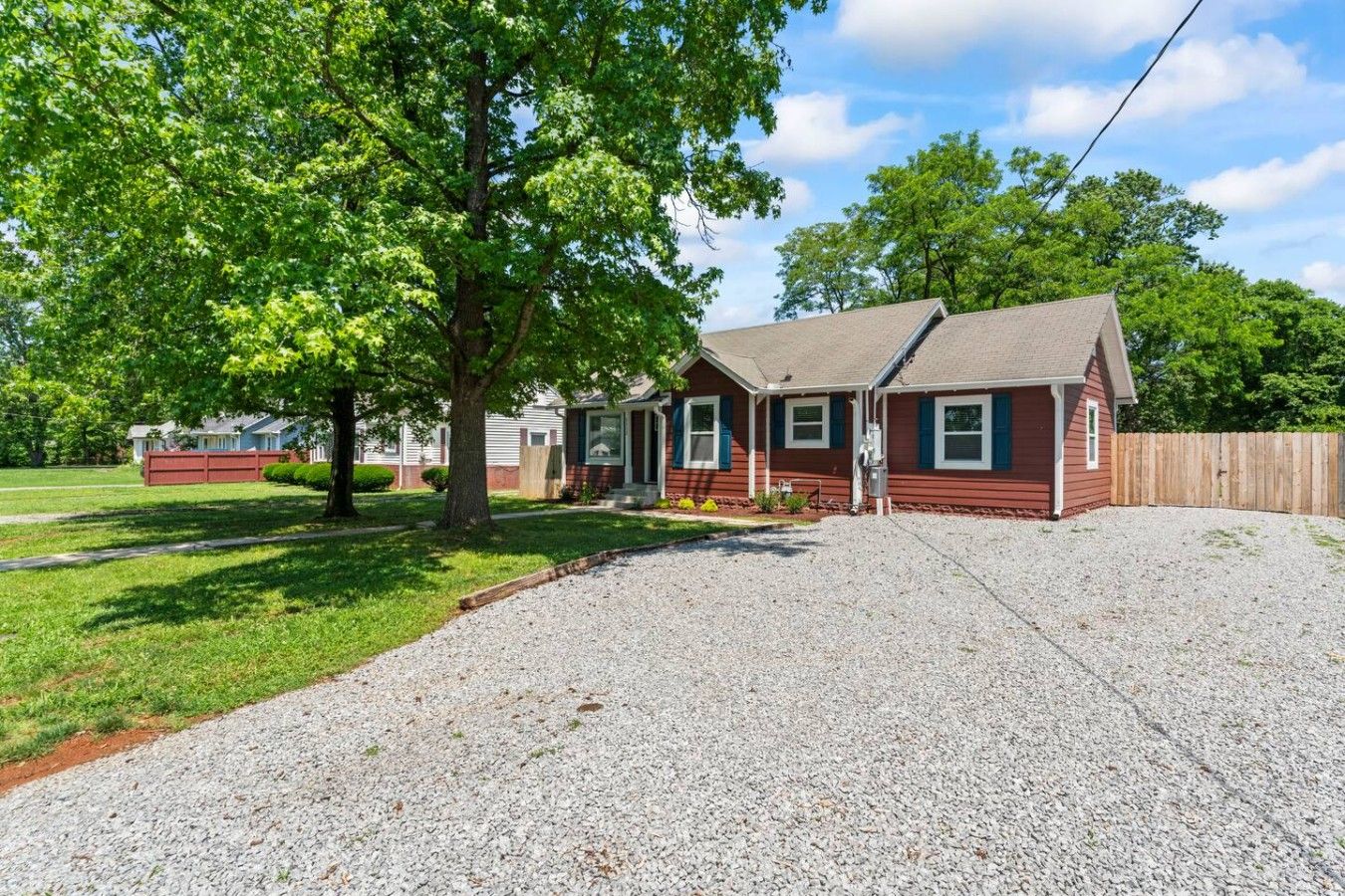 A single-story house with a gravel driveway, a wooden fence, and a large tree in front.