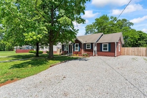 A single-story house with a gravel driveway, a wooden fence, and a large tree in front.