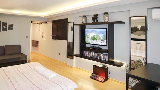 Ein modernes Hotelzimmer mit Bett, Wandfernseher, Bücherregal und Essecke mit Stühlen.