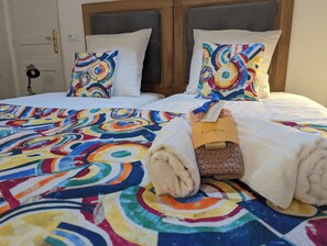 Habitación de hotel con una cama con colcha de diseño circular y colorido, dos almohadas con un diseño similar y una toalla doblada con una etiqueta que dice 'Bienvenue'.