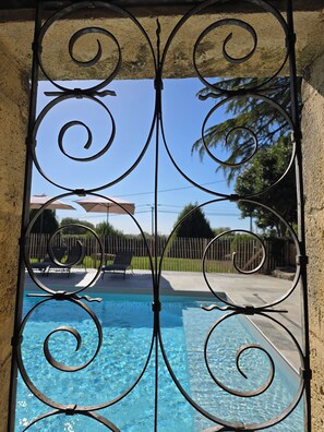 Una reja metálica con una piscina al fondo, cielo despejado y un patio trasero cercado con sillas y sombrillas.