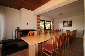 A dining area with a long wooden table, red cushioned chairs, a brick fireplace, and a stone wall.