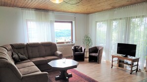 Sala de estar con sofá seccional, una mesa de centro, dos sillones, un televisor sobre un soporte de madera y vistas al exterior a través de una ventana.