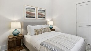 A neatly arranged bedroom with a bed, two bedside tables with lamps, and two framed artworks on the wall. - Bay St Louis Retreat | Downtown BSL (Bay St. Louis)