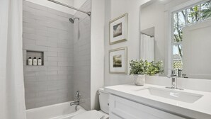 A modern bathroom with a white bathtub, toilet, and sink. There are framed pictures on the wall and a potted plant on the vanity.