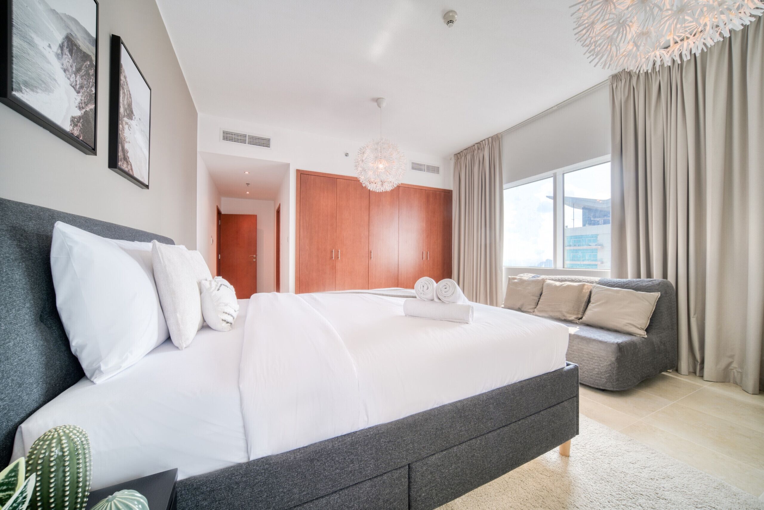 A modern hotel room with a large bed, a grey sofa, a cactus plant, and a view of the city.