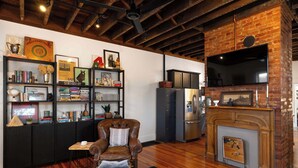 A cozy living room with a leather armchair, wooden coffee table, and a fireplace. There are bookshelves filled with books and decorative items, and a modern kitchen with a refrigerator and a flat-screen TV mounted on a brick wall. - 1BR Downtown, Close to Cincinnati Stadiums & Bars (Covington)