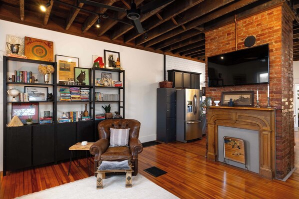 Een knus woongedeelte met een leren fauteuil, een houten salontafel en een open haard. Er staan boekenkasten vol boeken en decoratieve objecten, en een moderne keuken met een koelkast en een flatscreen televisie die aan een bakstenen muur is bevestigd.
