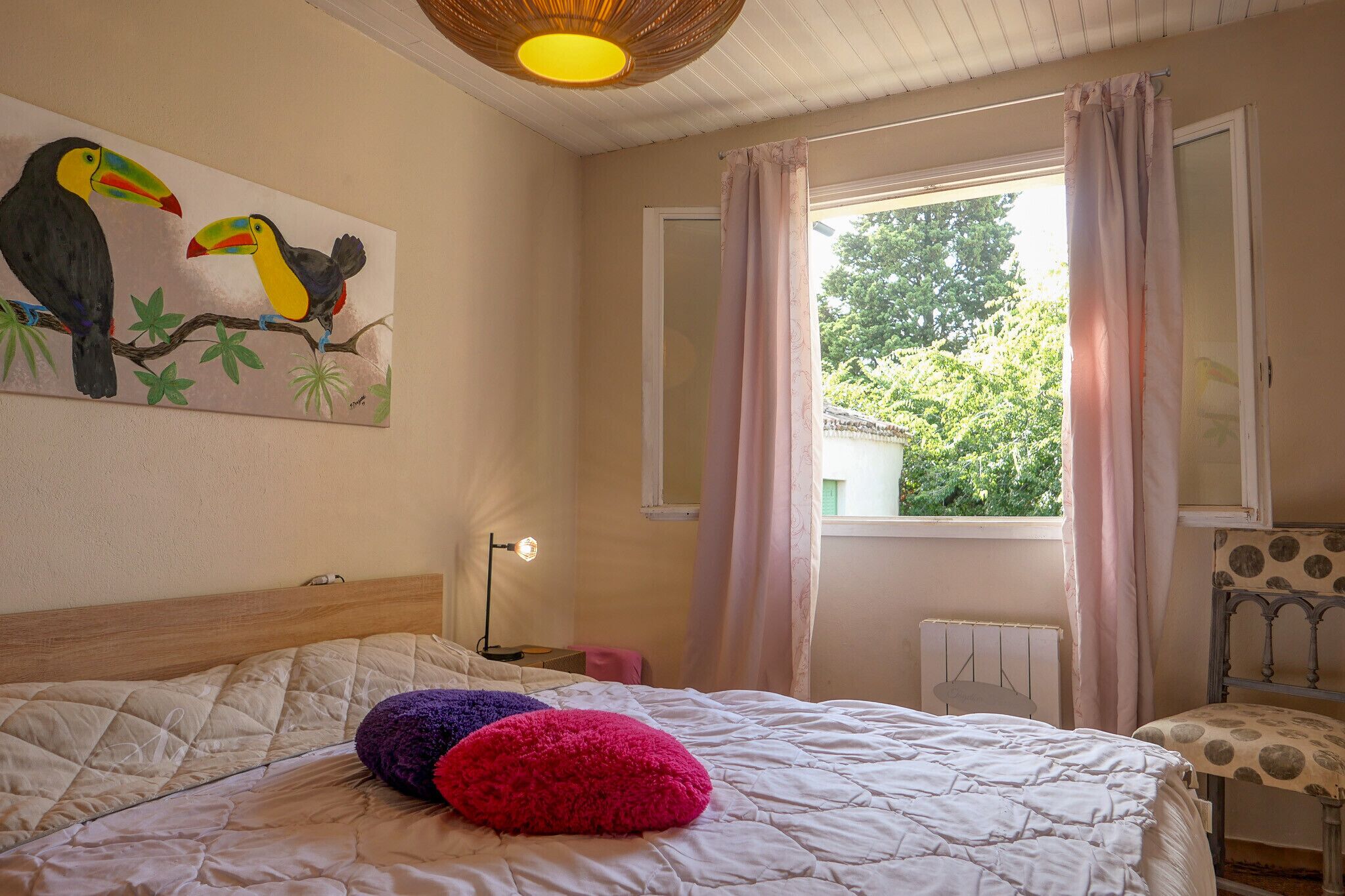Ein Schlafzimmer mit einem Bett, einem Papageienbild, einem Fenster mit Vorhängen, einer Heizung und einem Sessel.