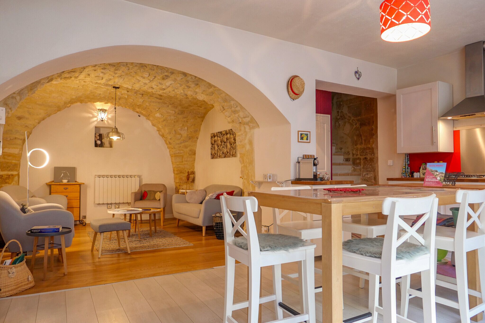 Una sala de estar acogedora con un arco de piedra, un comedor con sillas blancas y una cocina con detalles en rojo.