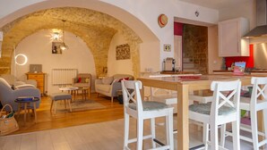 A cozy living room with a stone archway, a dining area with white chairs, and a kitchen with red accents. - Village house in the heart of Saint-Paul-Trois-Châteaux, Drôme Provençale (Saint-Paul-Trois-Châteaux)