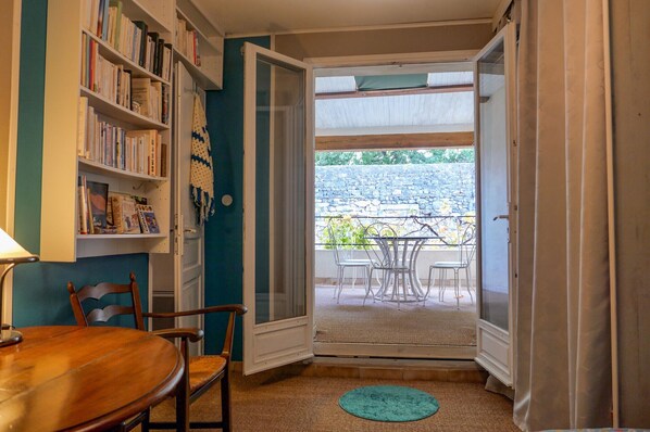 A room with a wooden dining table, chairs, a bookshelf, and a sliding glass door leading to an outdoor patio with a stone wall and metal chairs.