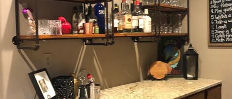 A bar area with wooden shelves holding bottles and glasses, a marble countertop, and a chalkboard menu.