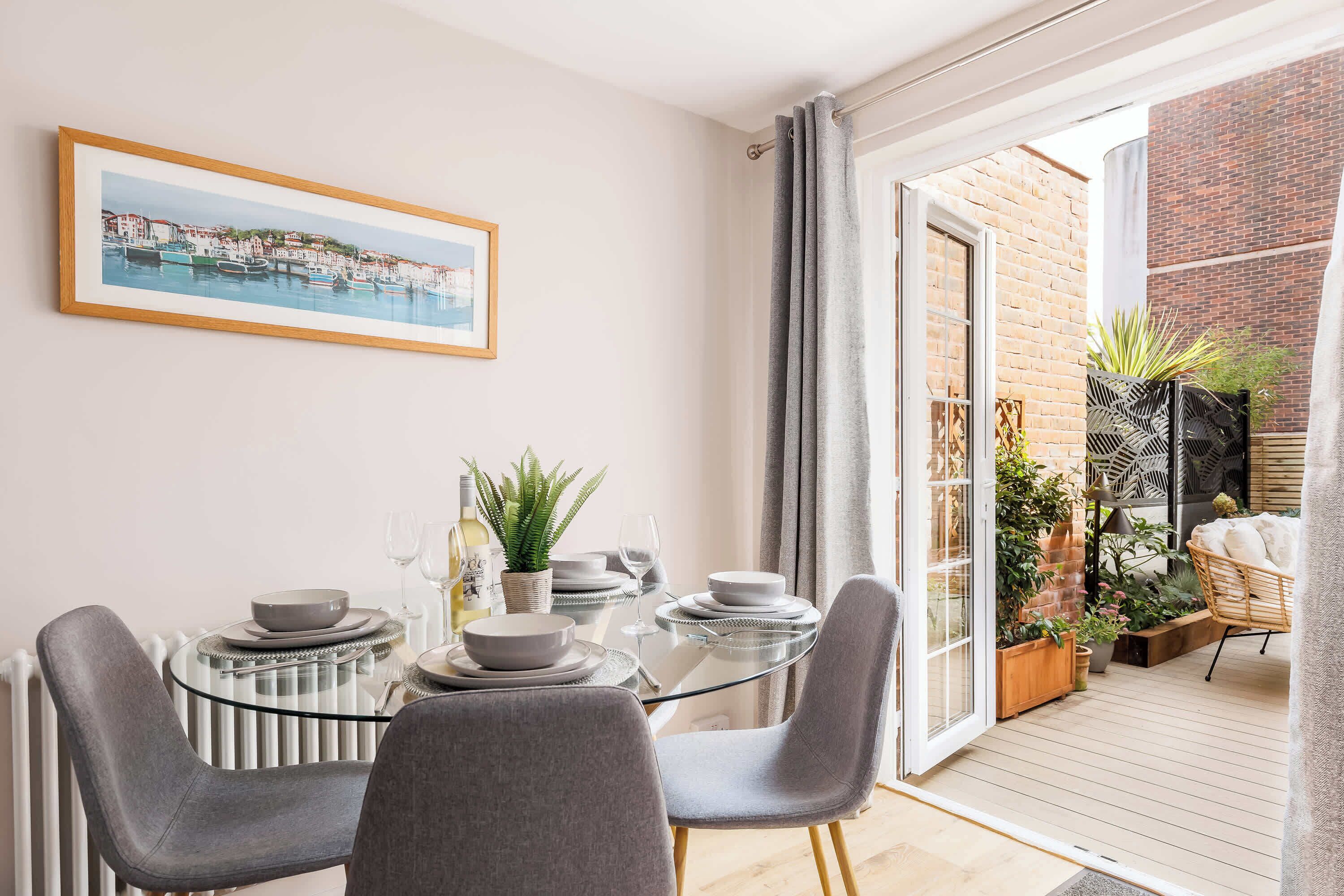 A dining area with a glass table, grey chairs, and a framed picture of a coastal town.