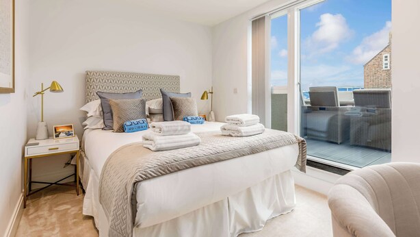 A bedroom with a large bed, a bedside table, a nightstand, a window with a view of outdoor furniture, and a headboard with a patterned design. - Vitamin Sea - Sun Terrace Overlooking Poole Quay (Poole)