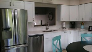 Cocina moderna con electrodomésticos de acero inoxidable, armarios blancos, encimera negra y sillas color turquesa.