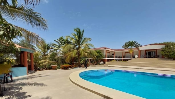 Une piscine à bord incurvé, entourée de palmiers et bordée d’un bâtiment au toit rouge.