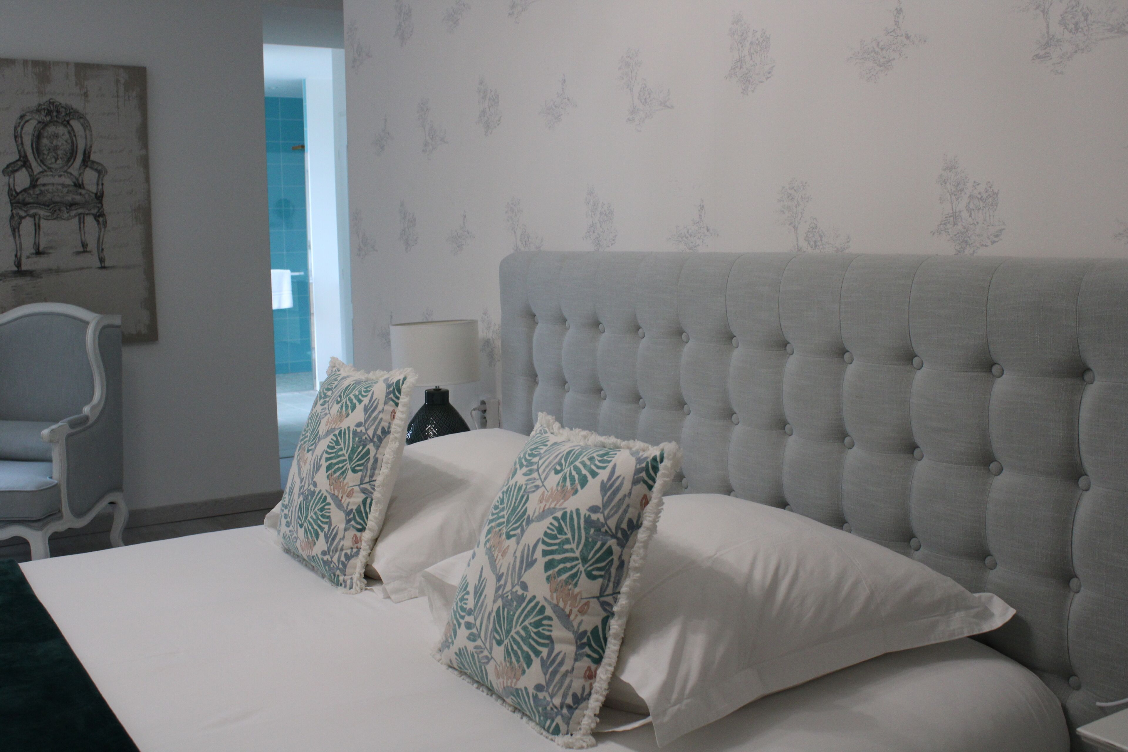 A neatly made bed with a tufted headboard, two decorative pillows, a white bedside table, and a chair.