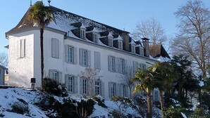 Um edifício grande e branco, com várias janelas e telhado inclinado, rodeado por neve no chão e árvores.