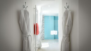 Um corredor com roupões brancos pendurados em ganchos, que leva a um banheiro com azulejos azuis e chuveiro.