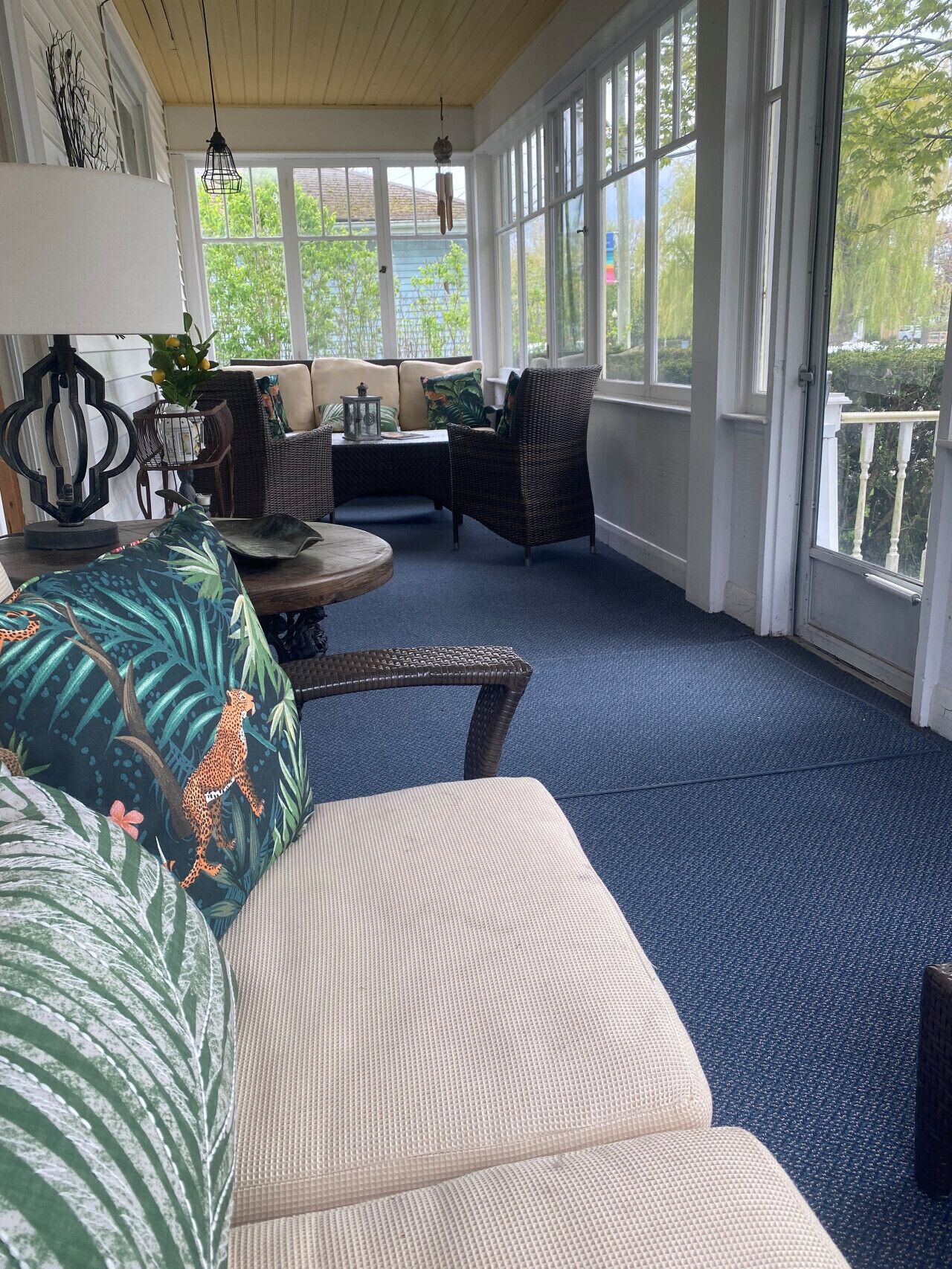 A sunlit room with a blue carpet, wicker furniture, and a view of greenery outside.