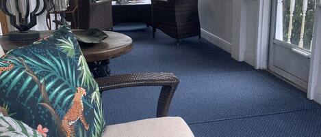 A sunlit room with a blue carpet, wicker furniture, and a view of greenery outside.