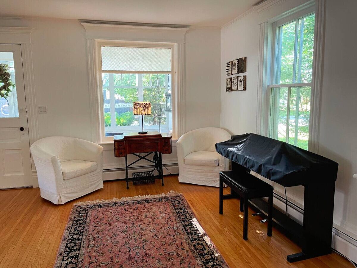 Un salon avec un piano, deux fauteuils blancs, une petite table en bois et une grande fenêtre donnant sur un paysage verdoyant.
