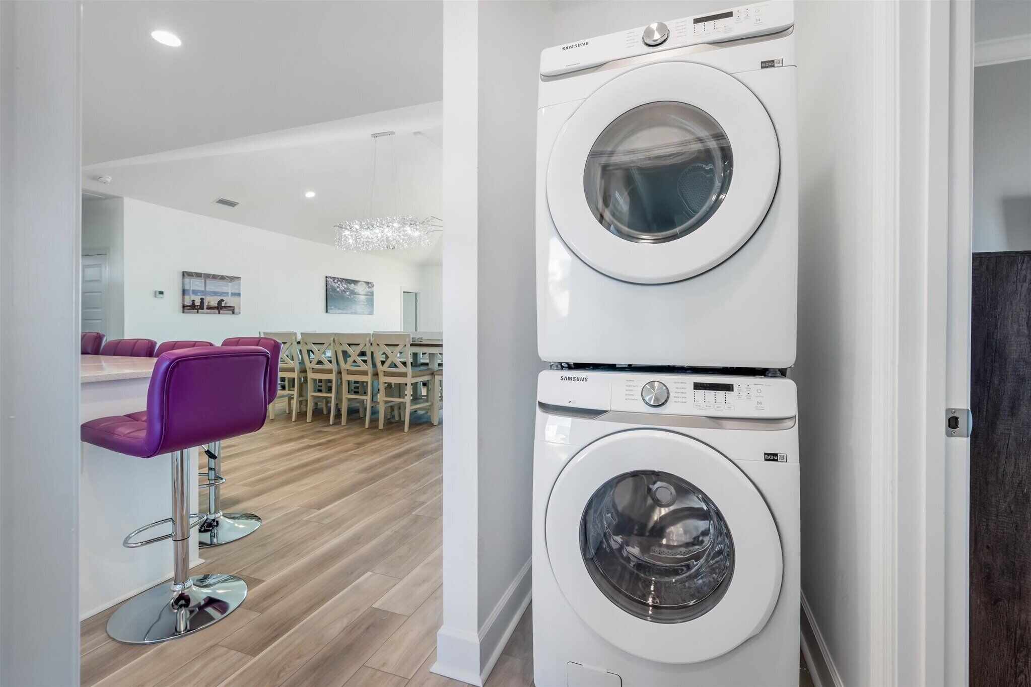 Une buanderie équipée de deux machines à laver Samsung superposées, d’un plancher en bois et d’un coin salon avec des chaises violettes.