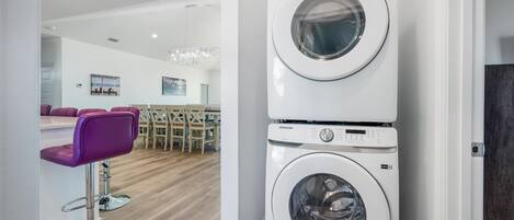 Une buanderie équipée de deux machines à laver Samsung superposées, d’un plancher en bois et d’un coin salon avec des chaises violettes.