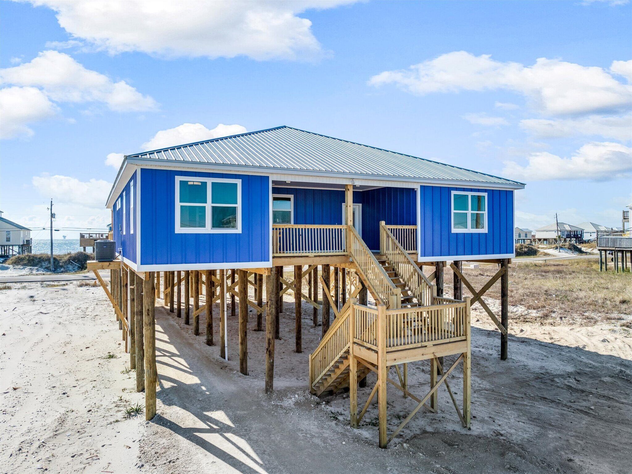 A blue beach house with a wooden deck and stairs, elevated on stilts.