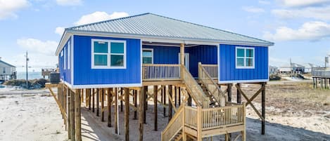 A blue beach house with a wooden deck and stairs, elevated on stilts.