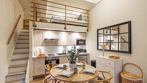 A compact kitchen with white cabinets, a microwave, and a sink. There is a dining table with four chairs and a potted plant. A staircase leads to a loft area with a bed. - Elegant flat with AC, heart of Nice (Nice)