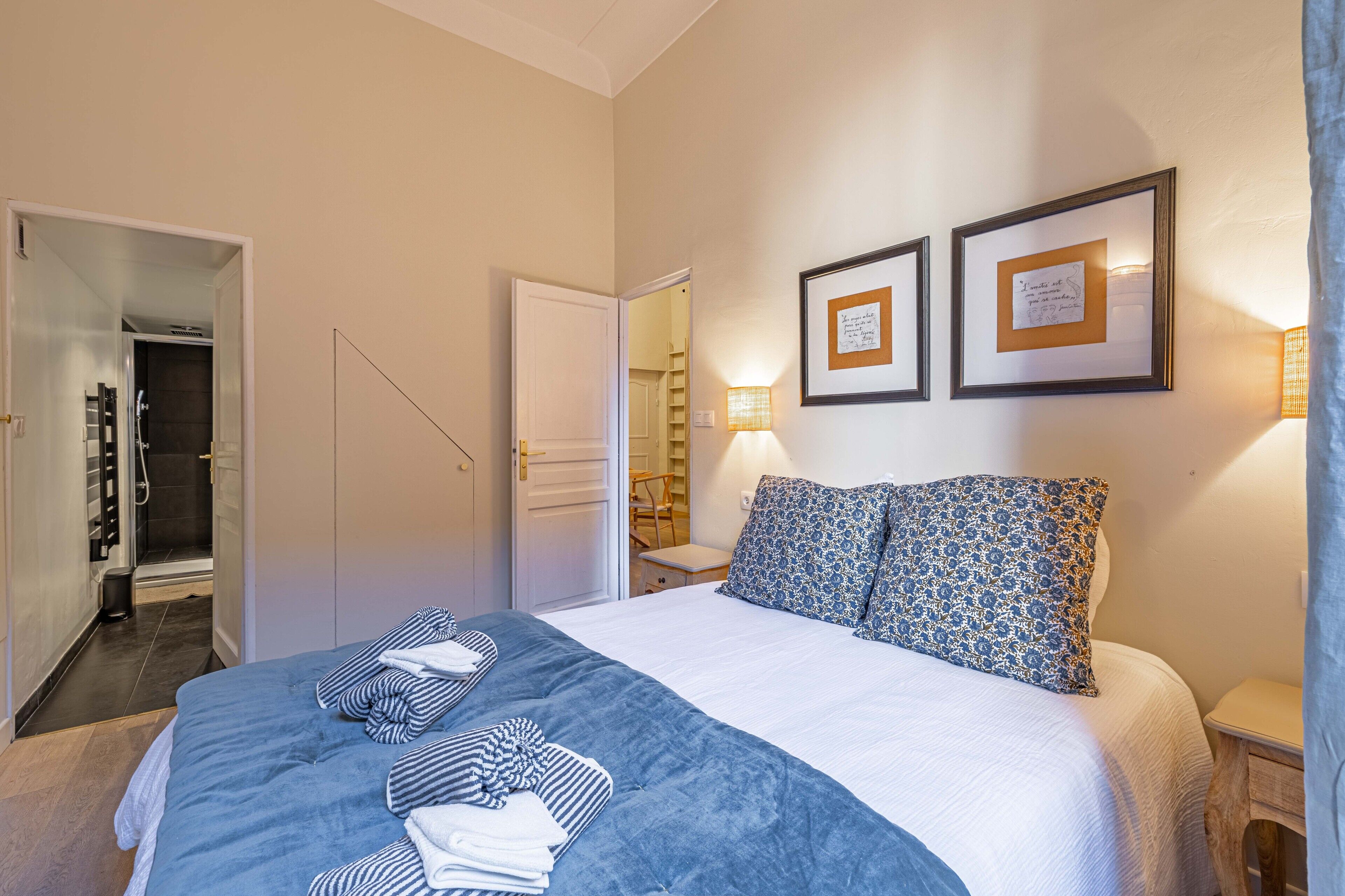Une chambre d’hôtel avec un lit, deux tableaux encadrés, une table de chevet et une salle de bain visible à travers une porte ouverte.