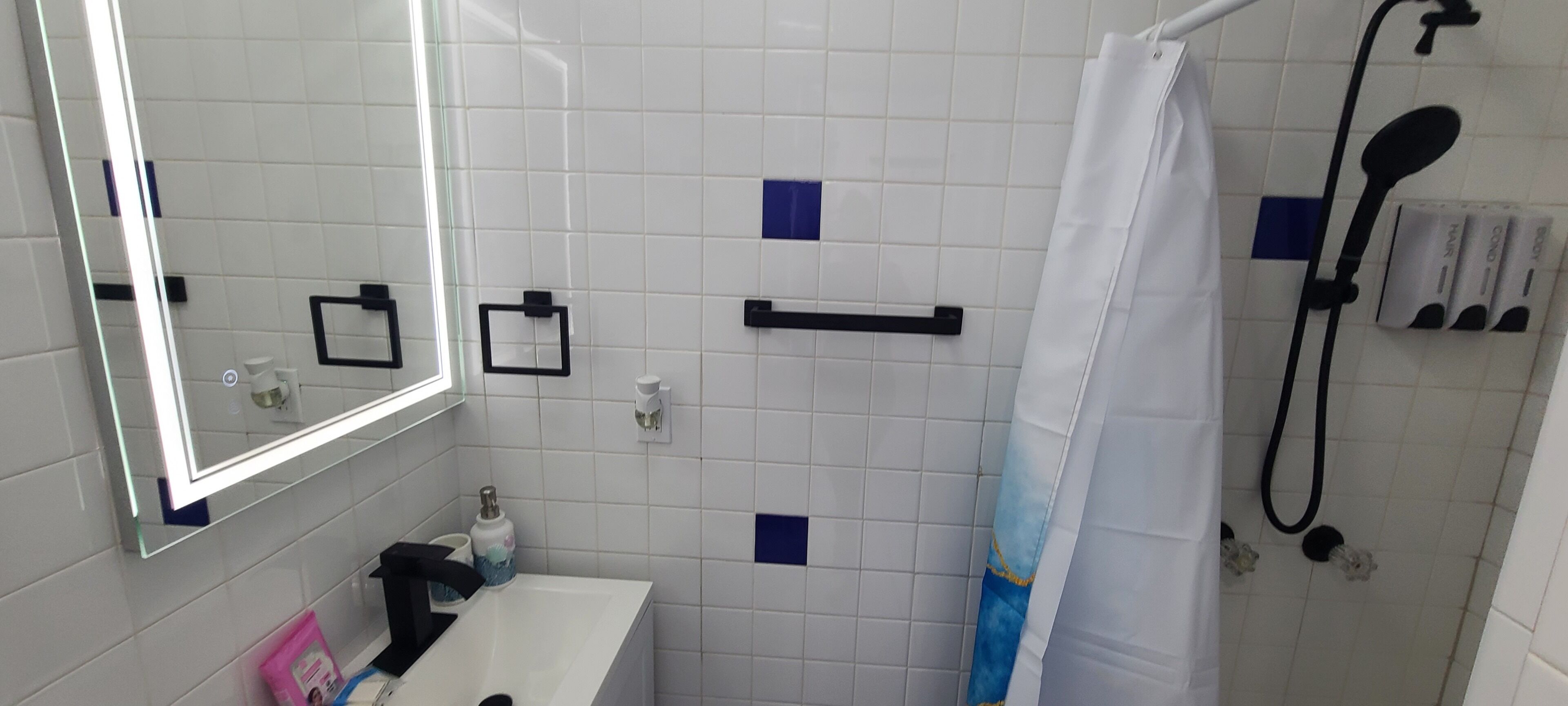 Ett badrum med vita kakelväggar, en spegel, ett handfat, en dusch och en handdukshängare.