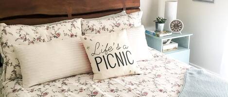 A bedroom with a floral bedspread, a pillow with "Life's a PICNIC" text, a wooden headboard, a small bedside table with a plant, and a framed picture on the wall.