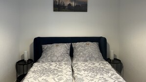 Un lit bien fait, avec une literie à motifs floraux, deux tables de chevet ornées de bougies et un tableau représentant un paysage accroché au mur.