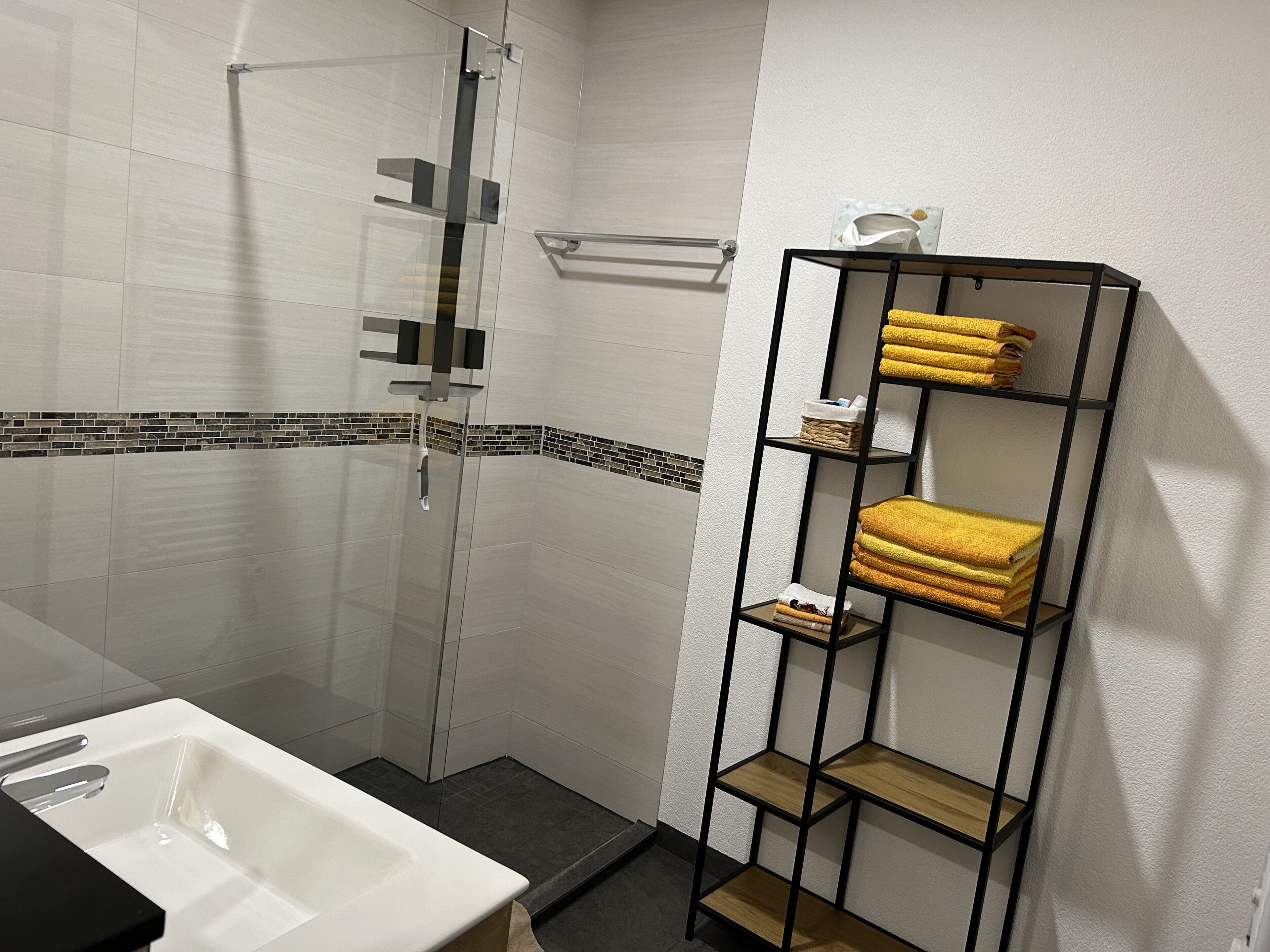 A bathroom with a glass shower enclosure, a white sink, and a black metal shelf with towels.