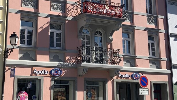 Un bâtiment rose dont la façade porte l’inscription « Apollo », avec un balcon orné de balustrades en fer forgé et des plantes en pot à l’entrée.