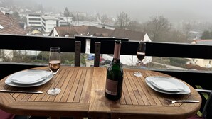 Une table en bois, dressée pour deux personnes, avec des assiettes, des verres et une bouteille de vin, donnant sur un quartier résidentiel plongé dans la brume.