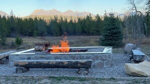 Un foyer en pierre avec des bûches et un feu allumé, entouré de rochers et d’arbres, avec des montagnes en arrière-plan.