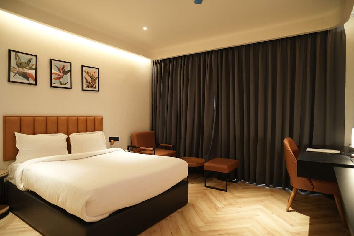 Een moderne hotelkamer met een groot bed, twee stoelen, een bureau en een groot raam met donkere gordijnen.