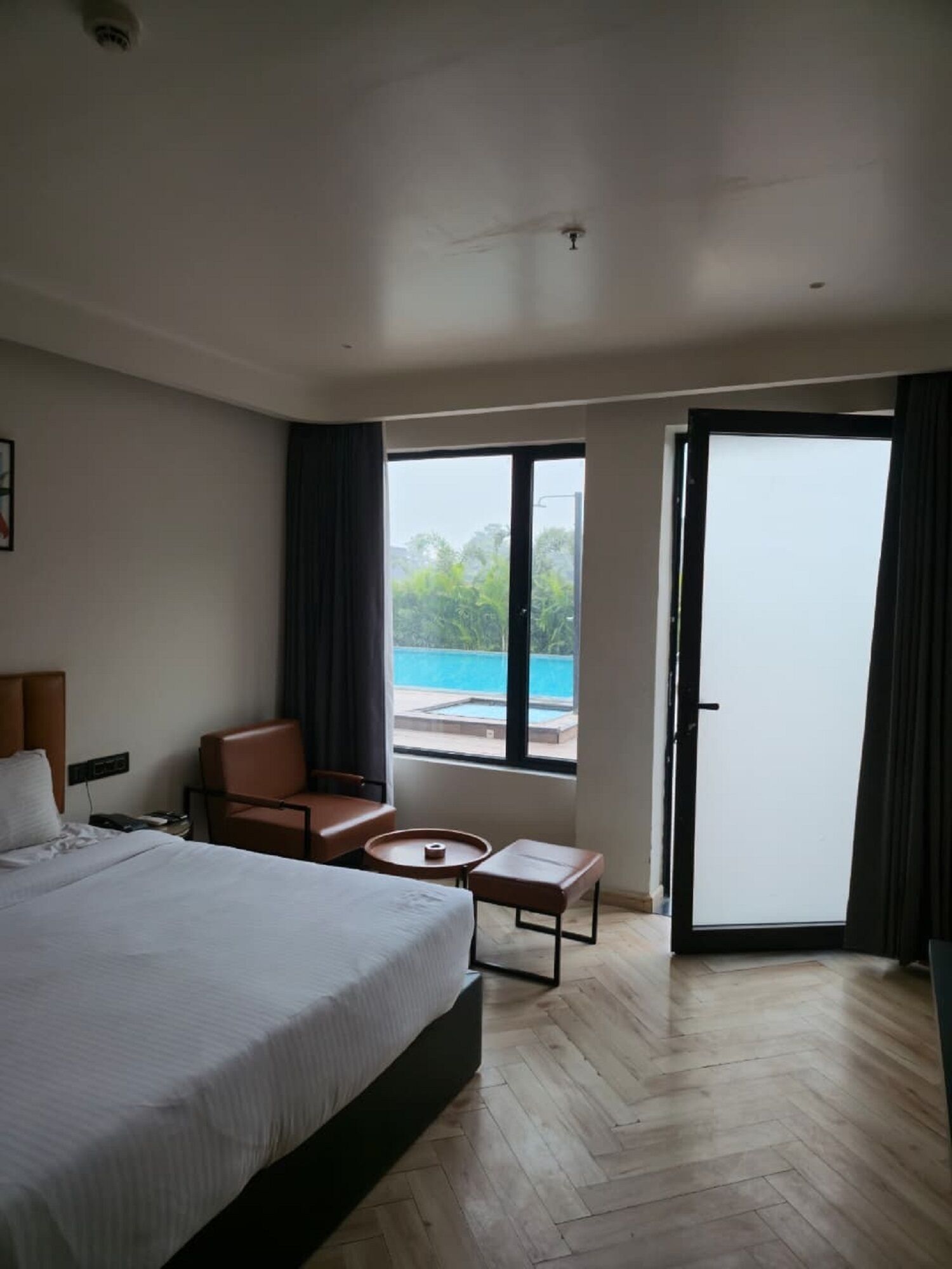 Ein Hotelzimmer mit einem großen Bett, einer Sitzecke mit Tisch und Blick auf einen Swimmingpool durch die Schiebetür.