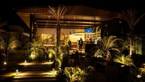 Un área de comedor al aire libre con mesas y sillas, rodeada de palmeras y plantas, bajo una estructura cubierta con un bar visible al fondo.