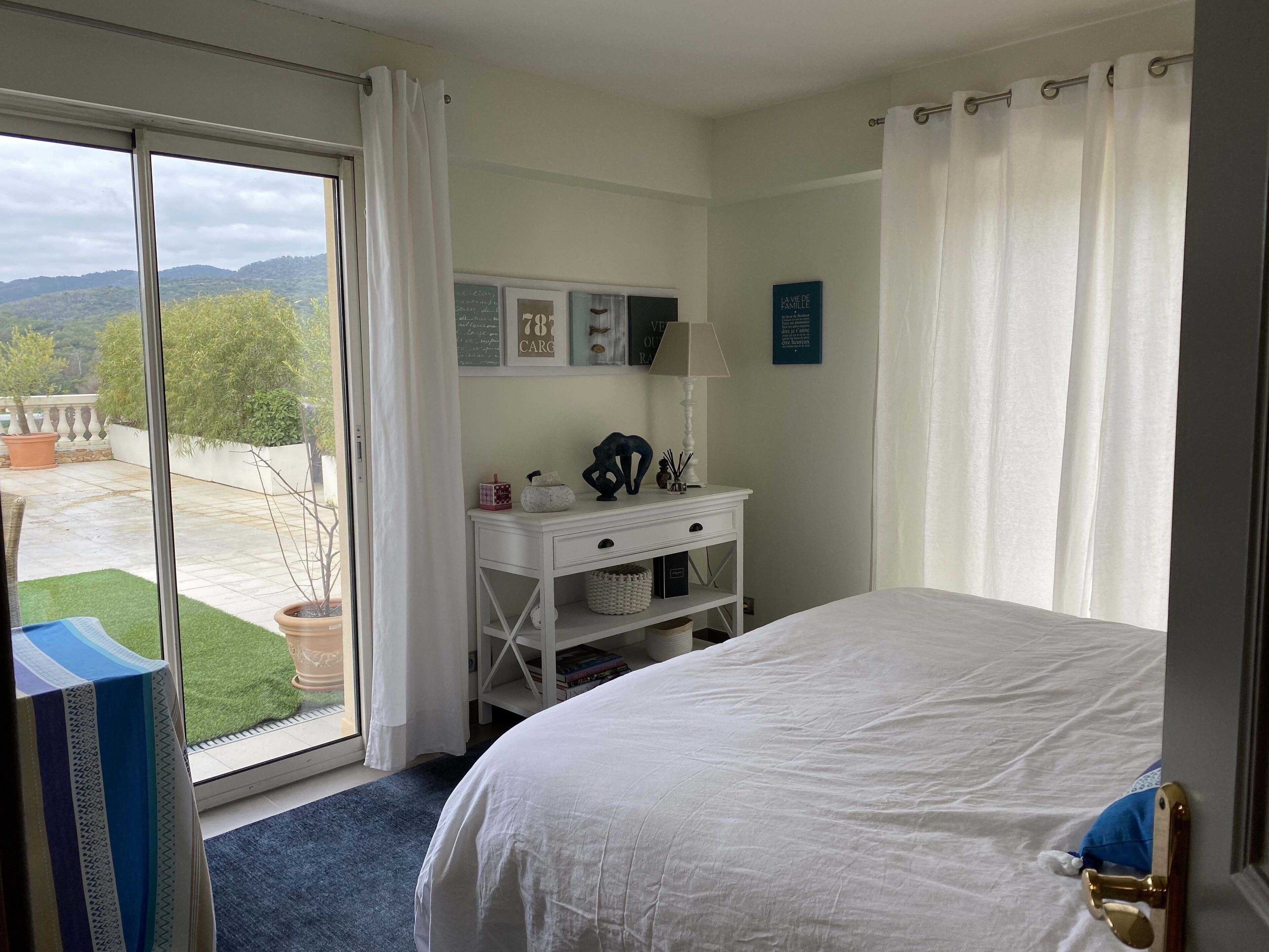 A bedroom with a large bed, a bedside table with a lamp, a window with curtains, and a view of the outdoors.