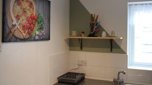 A kitchen with a painting of food, a sink, a window, and a shelf with kitchen utensils. - Apartment - Sleeps 4 - Garden - Parking (Romford)