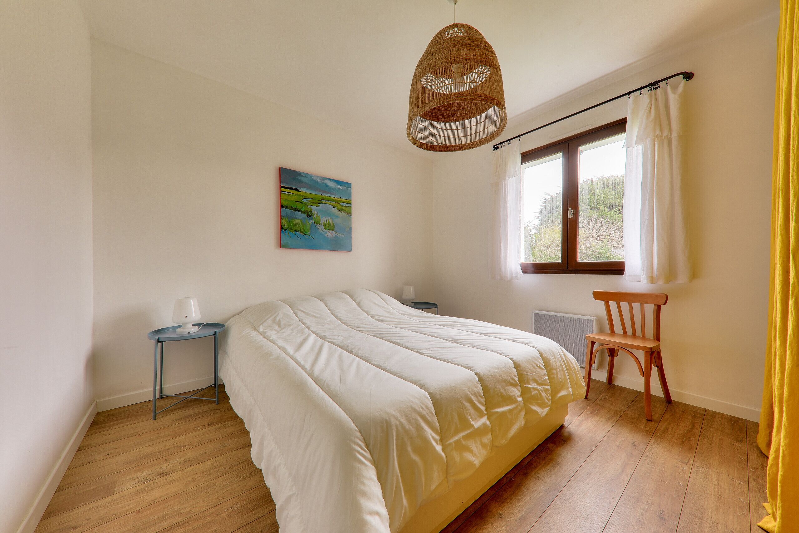 A bedroom with a bed, bedside table, lamp, chair, window, and artwork.