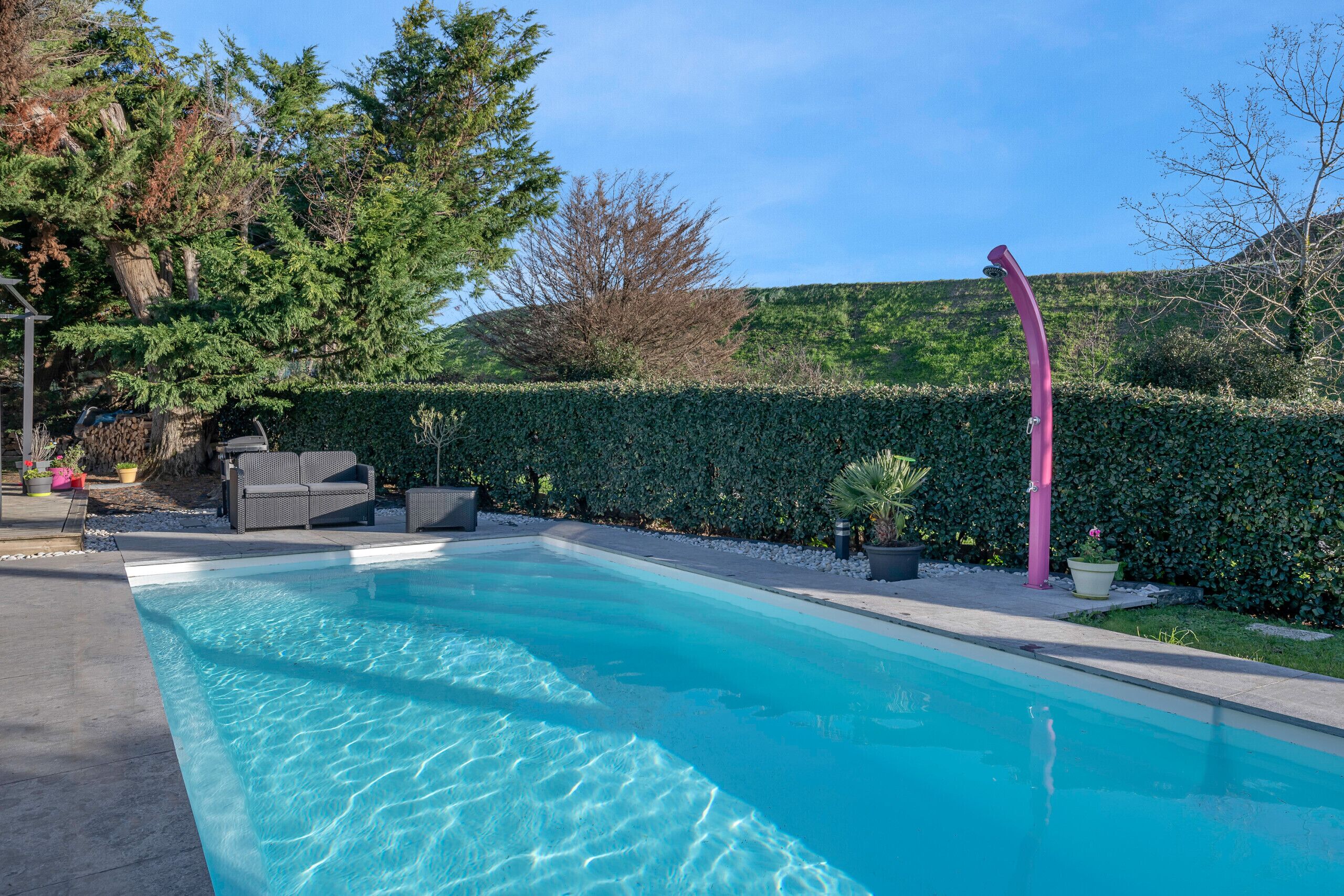 Uma piscina retangular com água azul cristalina, rodeada por um viveiro, um poste rosa e mobiliário de exterior.