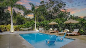 Une piscine rectangulaire bordée de chaises longues et d’ombrelles, entourée de palmiers et de plantes tropicales.